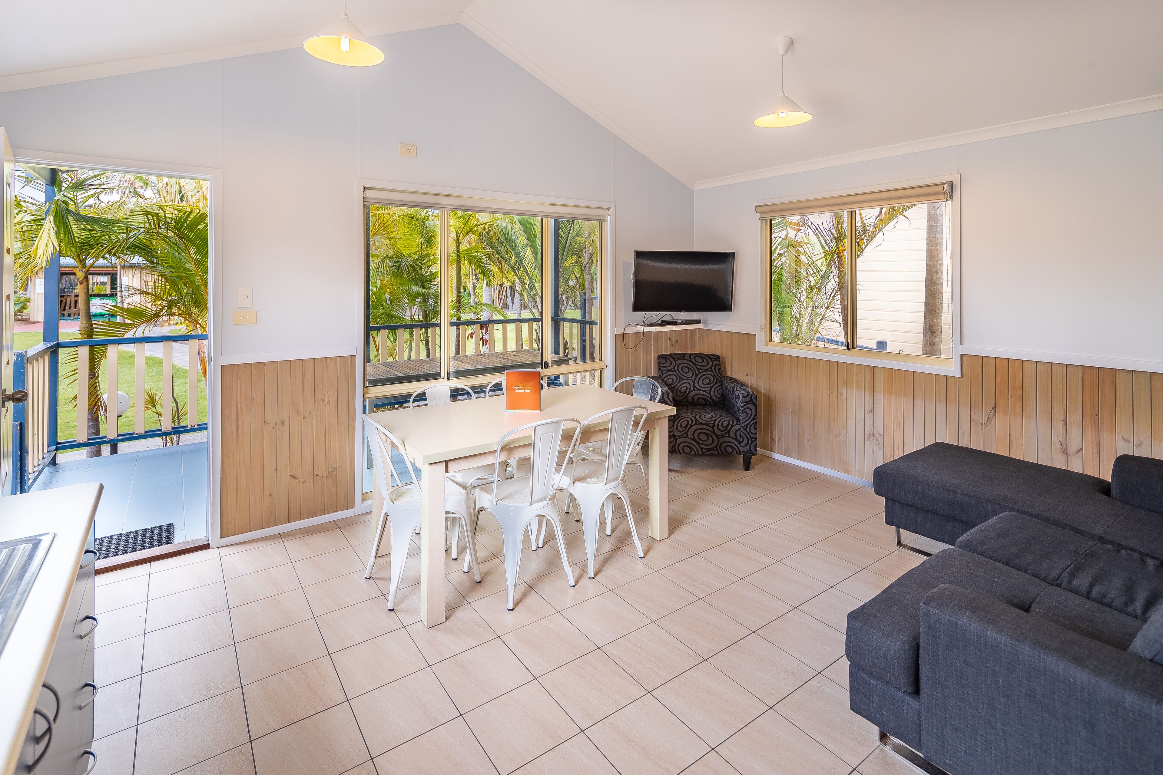A spacious room with a dining table and chairs, a sofa, a TV, and large windows offering a view of palm trees.