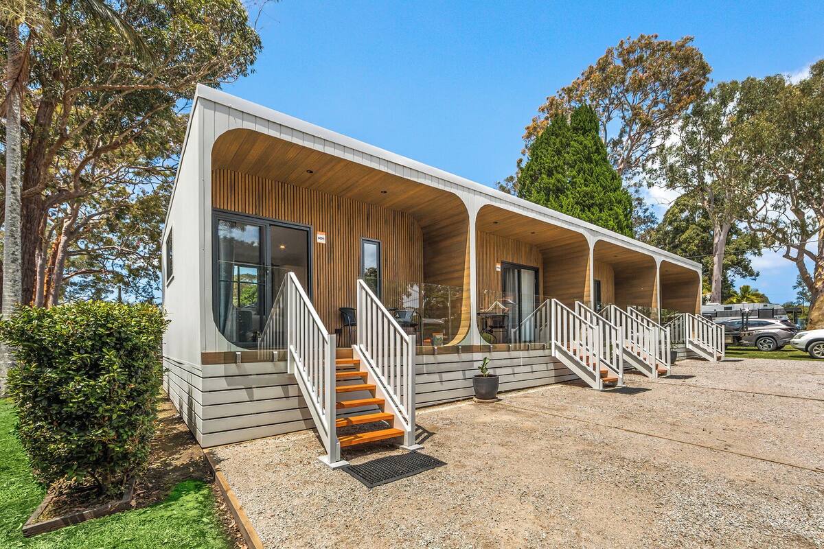 A modern caravan-style building with multiple covered verandas, a staircase leading to the entrance, and a gravel driveway.