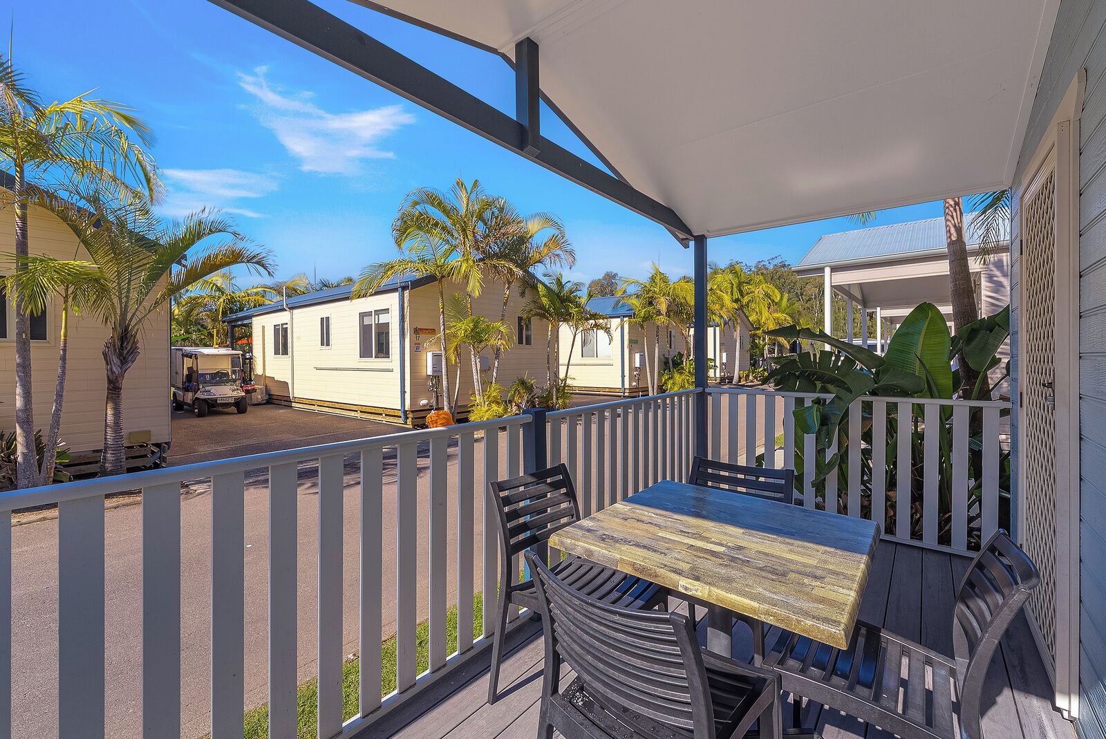 A covered outdoor area with a table and chairs, a white fence, and a view of mobile homes in the background.
