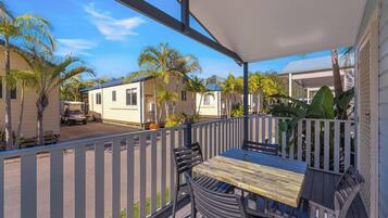 A covered outdoor area with a table and chairs, a white fence, and a view of mobile homes in the background.