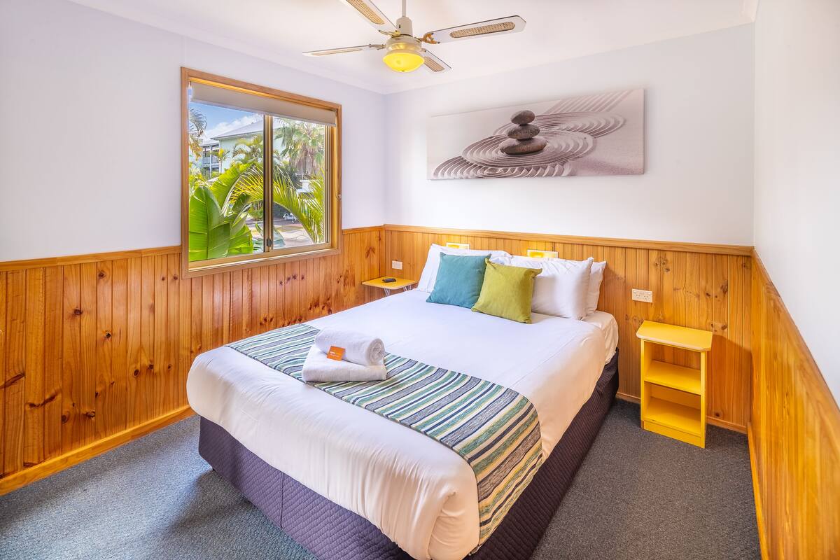 A hotel room with a bed, wooden paneling, a window with a view, a ceiling fan, and a wall-mounted artwork.