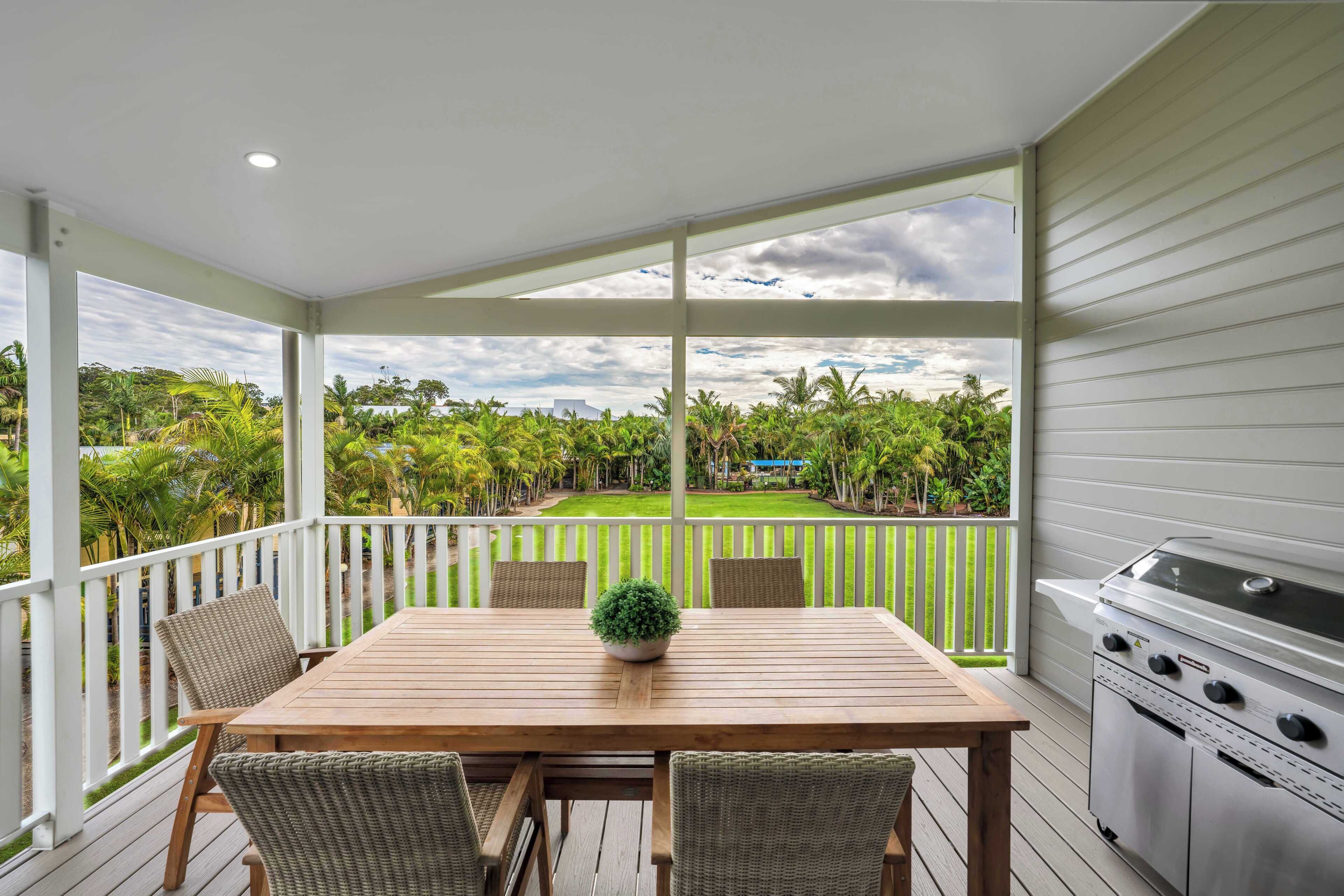 A covered outdoor patio with a wooden table and chairs, a grill, and a view of a lush garden.