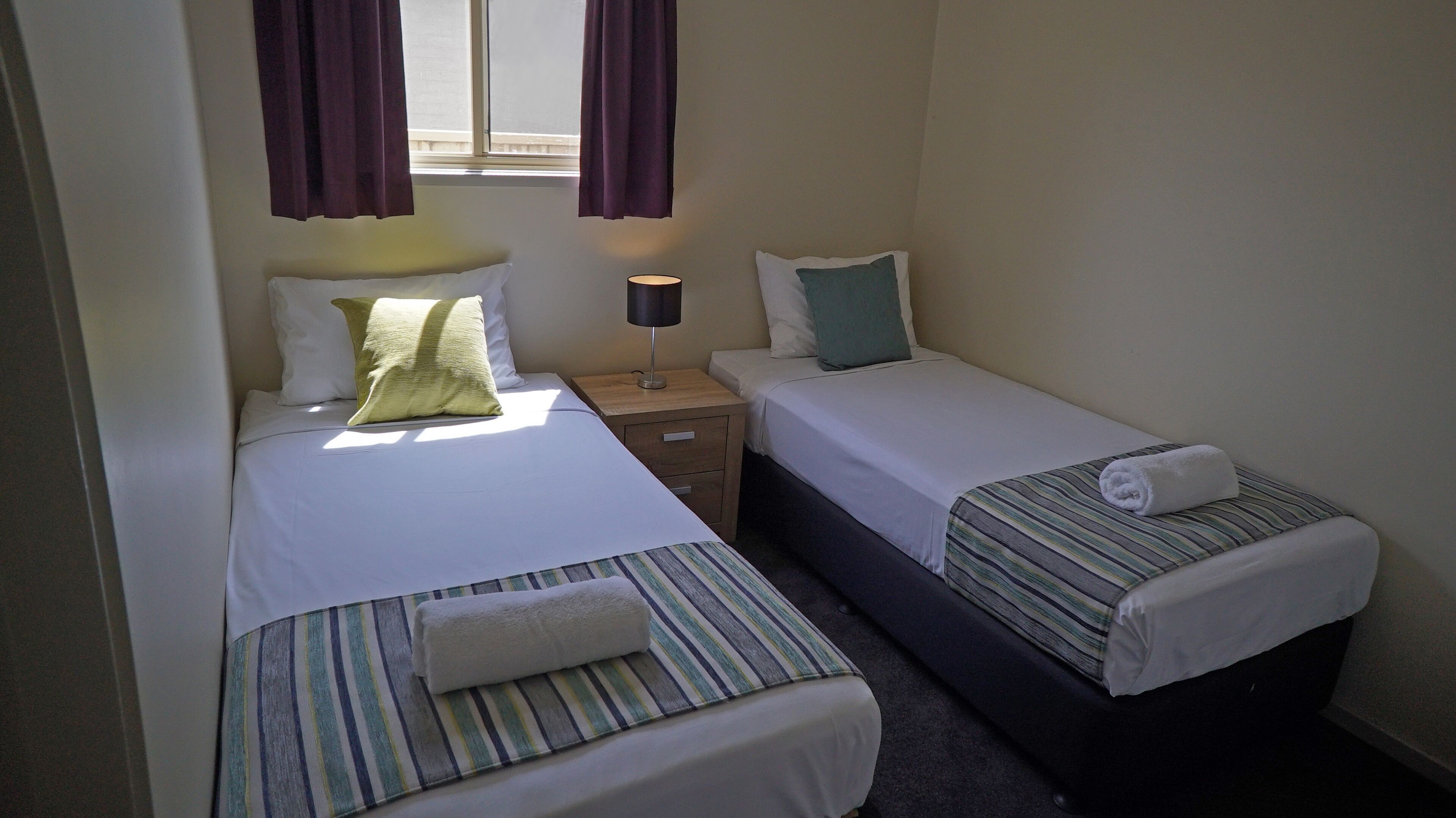 A hotel room with two single beds, a nightstand with a lamp, and a window with curtains.