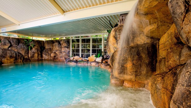 A pool with a waterfall feature surrounded by large rocks and a glass enclosure in the background.