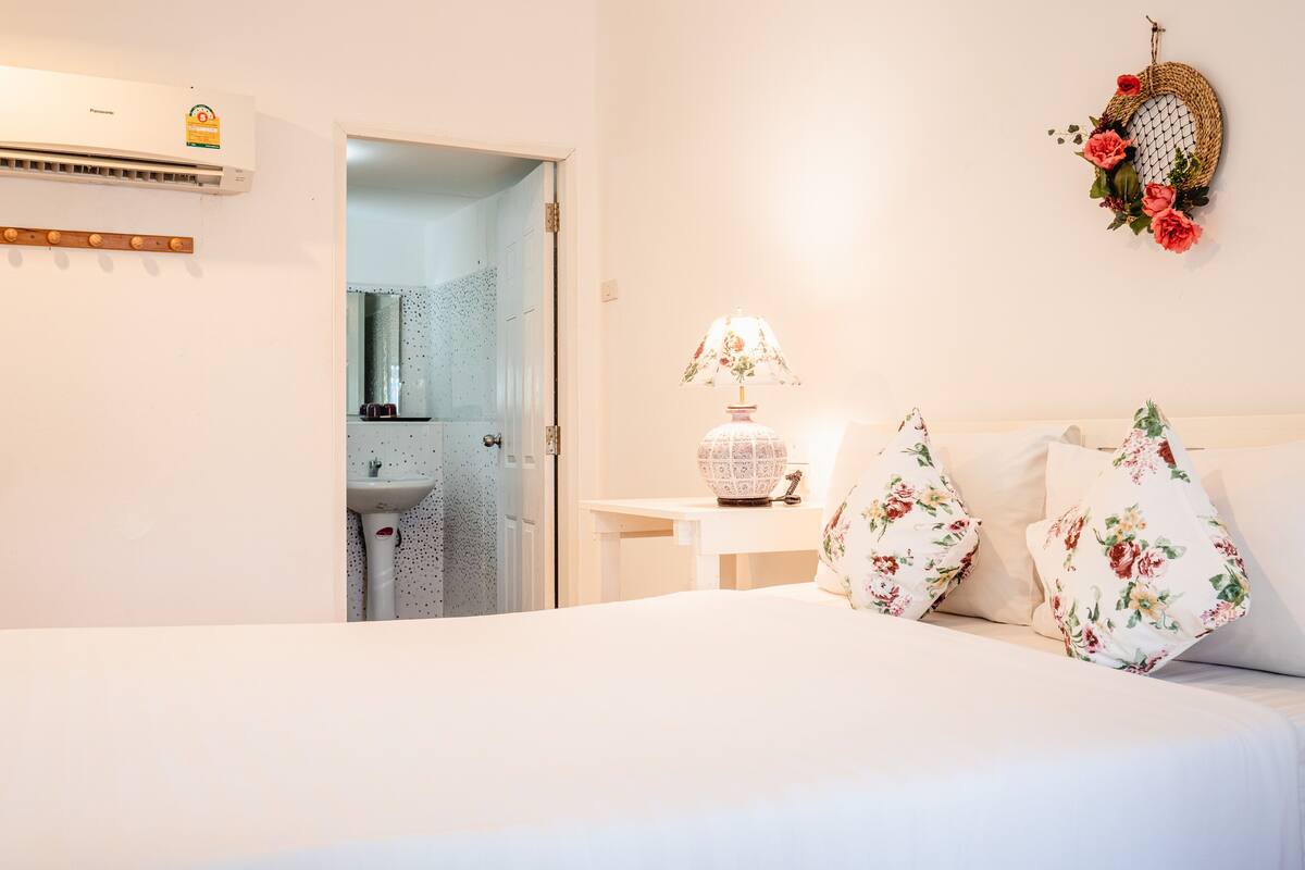 A bedroom with a bed, a bedside table with a lamp, a wall-mounted air conditioner, and a decorative wall hanging.
