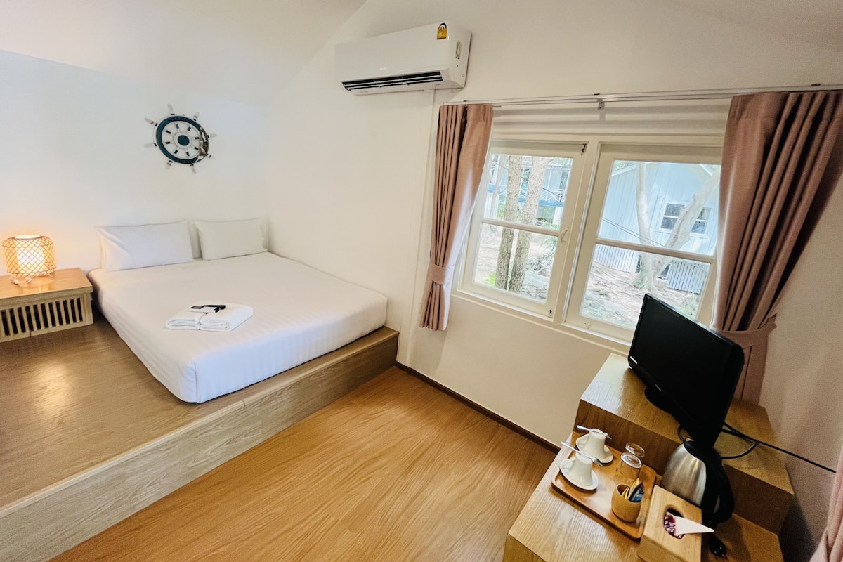 A hotel room with a bed, a desk with a computer, a window with curtains, an air conditioner, and a wall clock.