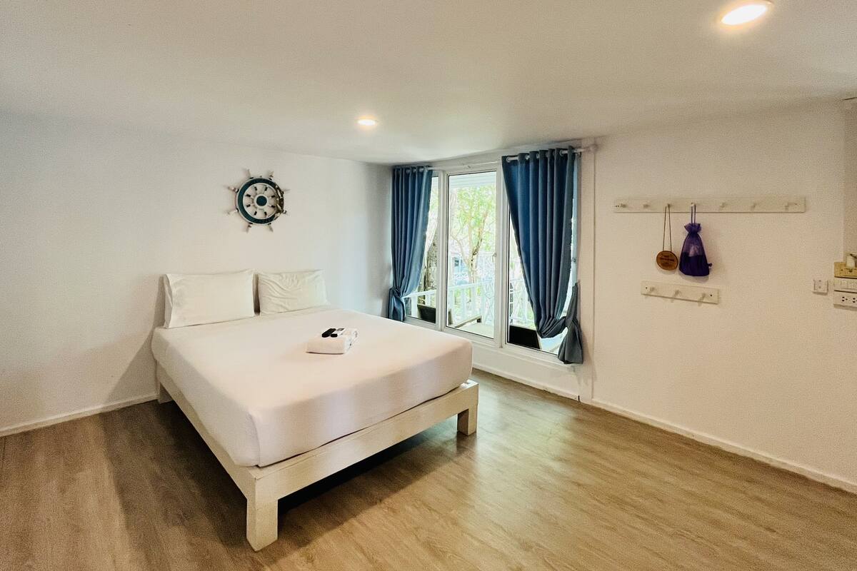 A bedroom with a bed, a window with curtains, a wall clock, and a small shelf with items.