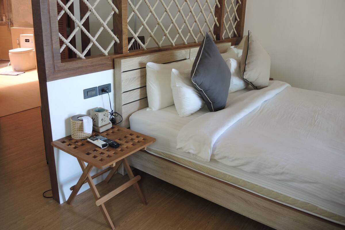 A neatly made bed with white linens, a wooden nightstand with a remote control, and a small table with a cup and tissues.