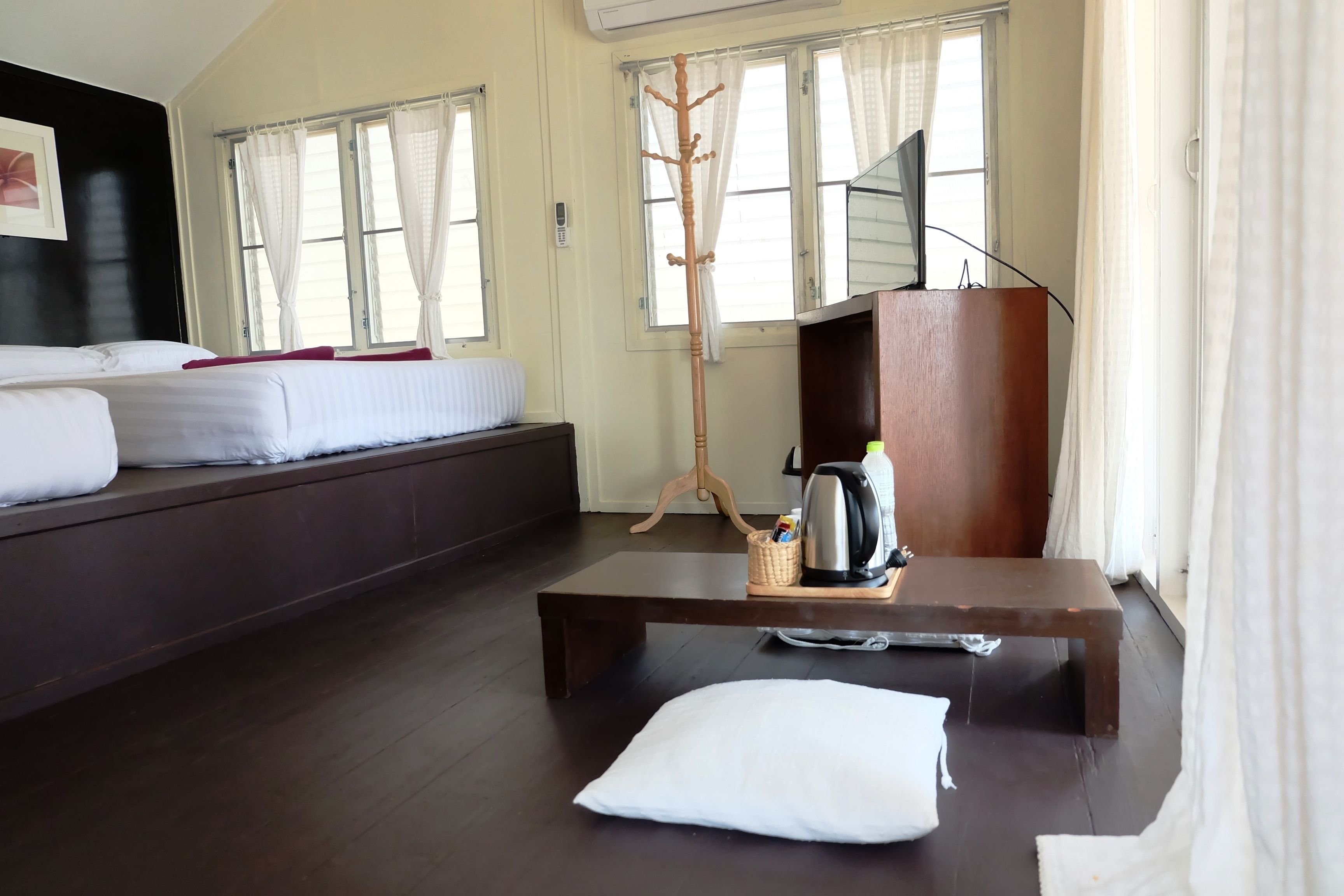 A hotel room with a bed, a wooden coffee table with a kettle and a basket, a coat rack, and a TV on a wooden stand.