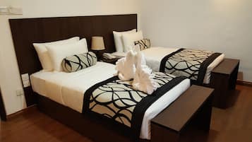 A double bed with a dark headboard, white bedding, and decorative towels. There is a bedside table with a lamp and a phone. The room has wooden flooring and a plain wall.