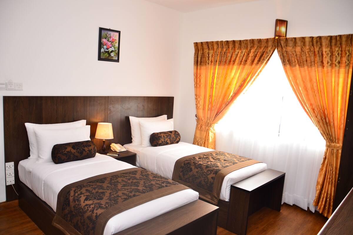 A hotel room with two beds, a nightstand with a lamp, a telephone, and a window with curtains.