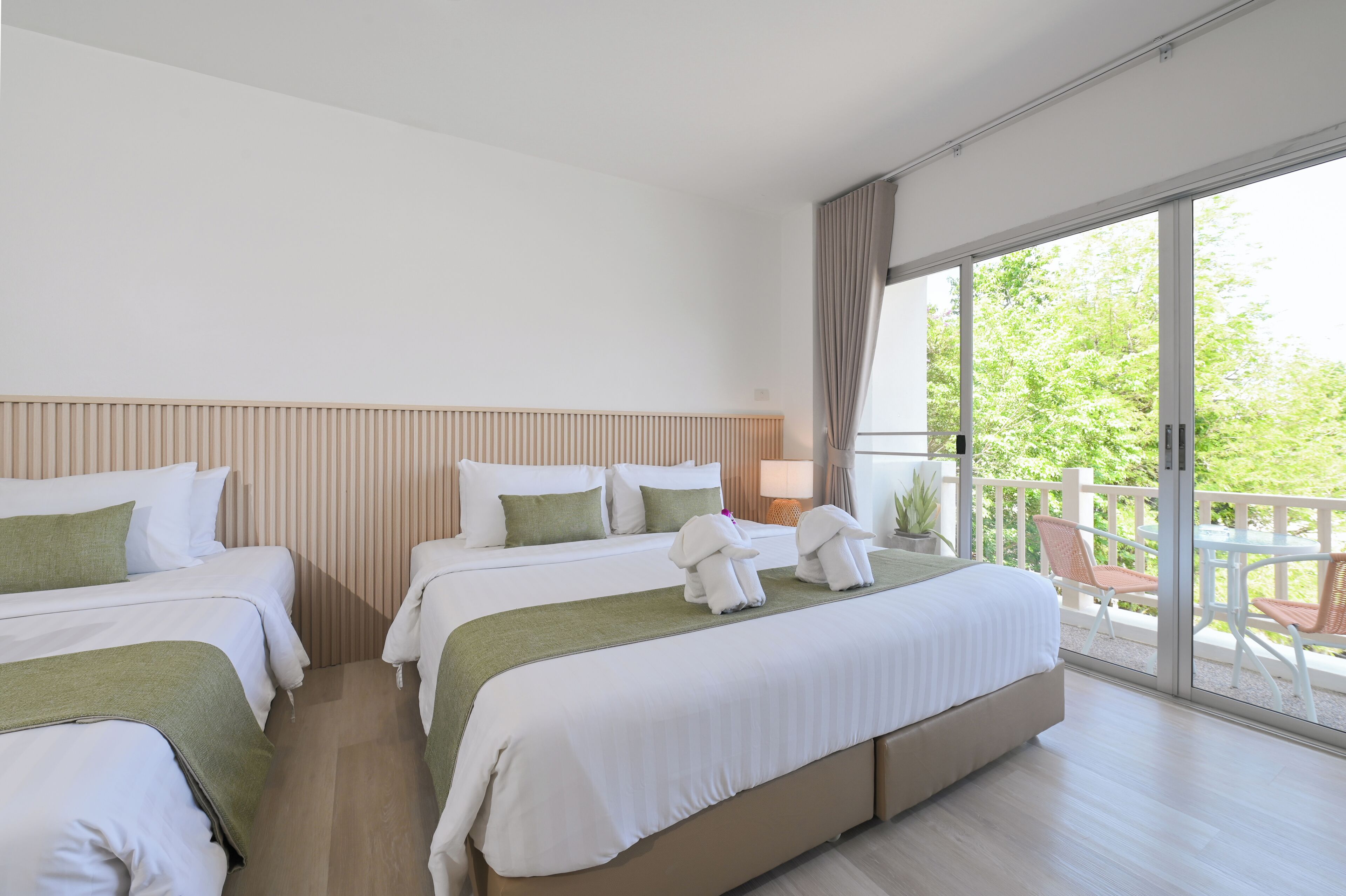 A hotel room with two beds, a balcony with chairs, and a view of greenery.