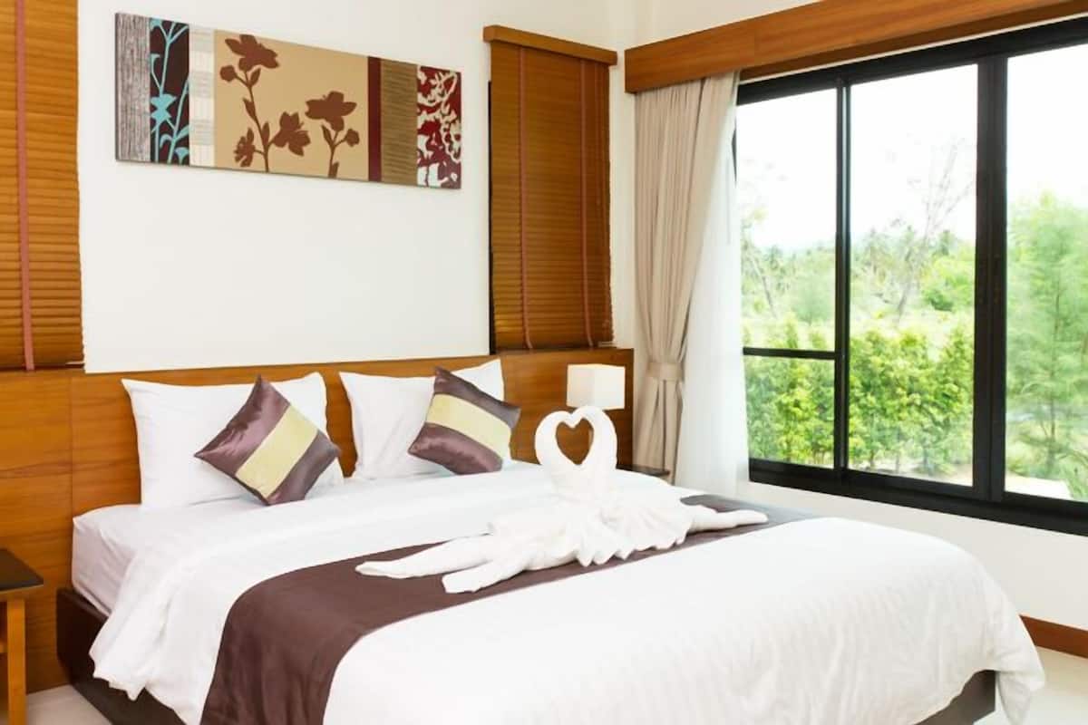 A neatly made bed with a brown bedspread and white pillows, a bedside table with a lamp, and a large window with a view of greenery.