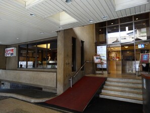 Entrada de hotel con una rampa que lleva a las escaleras, un mostrador de recepción y un letrero visible del restaurante.
