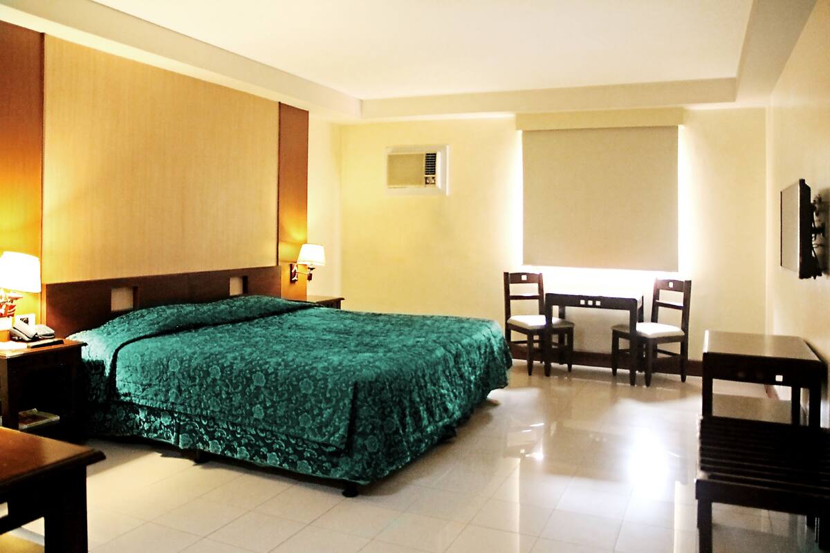 A hotel room with a large bed, two chairs, a small table, a nightstand, and a window with blinds.