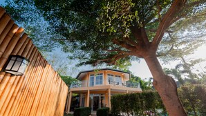 Een twee verdiepingen tellend huis met een balkon, omringd door bomen en een bamboescherm.