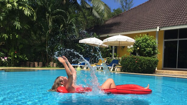 Una piscina con una persona acostada en un flotador rojo, rodeada de abundante vegetación y un edificio con tejado de baldosas.