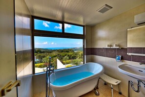 Une salle de bain équipée d’une baignoire, d’un lavabo et d’une fenêtre donnant sur un paysage verdoyant et une vue lointaine.