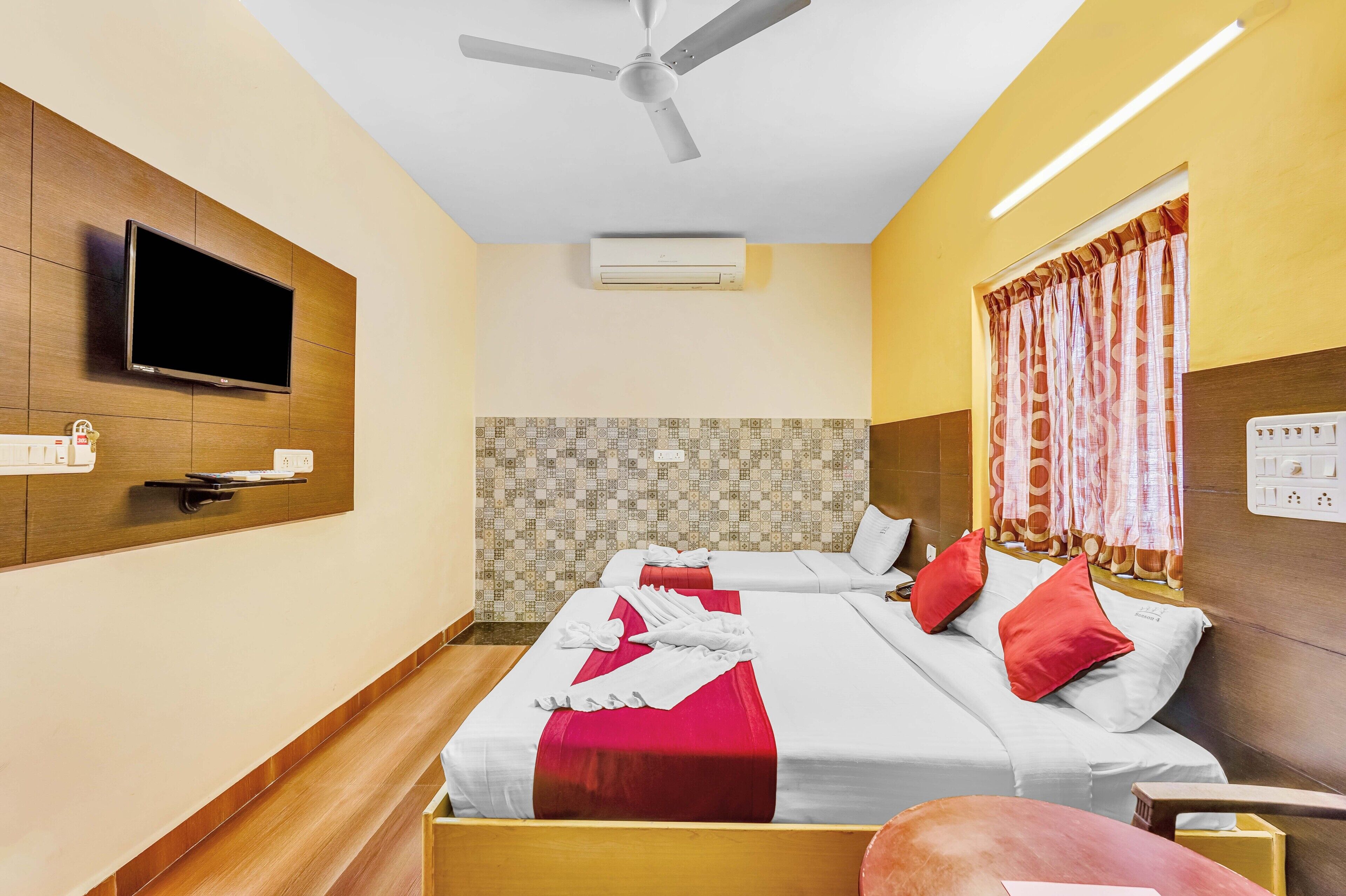 A hotel room with two beds, a TV, ceiling fan, air conditioning unit, and a window with curtains.