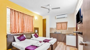 A hotel room with two beds, a ceiling fan, an air conditioning unit, a small kitchenette, and a TV mounted on the wall.