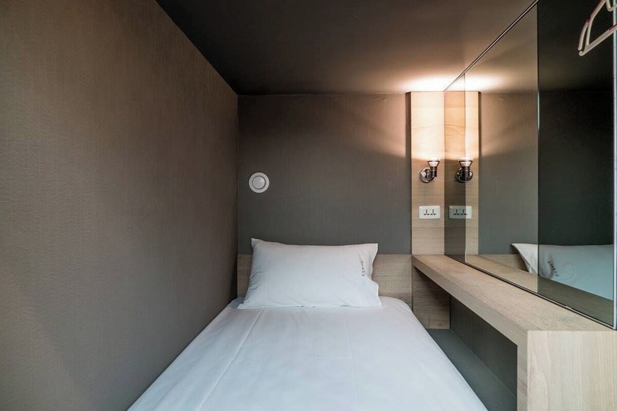 Una habitación de hotel moderna con una cama individual, un banco de madera, un espejo y una lámpara de pared.