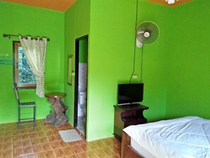 Une pièce aux murs verts, avec un lit, un téléviseur posé sur un meuble, un ventilateur, une petite table, une chaise et une fenêtre avec des rideaux.