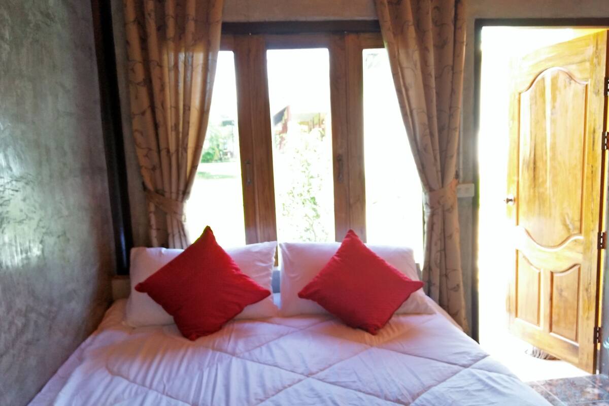 A bed with white bedding and two red pillows, a wooden door, and a view of greenery outside.
