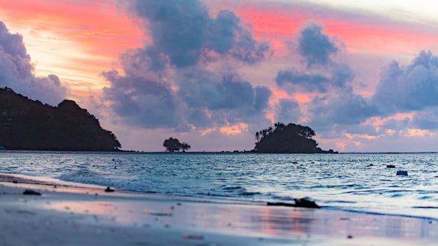 Una playa con aguas tranquilas, formaciones terrestres a lo lejos y un cielo al atardecer.