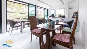 Un comedor espacioso con mesas y sillas de madera, ventanas grandes con marcos verdes y vistas a la vegetación exterior.