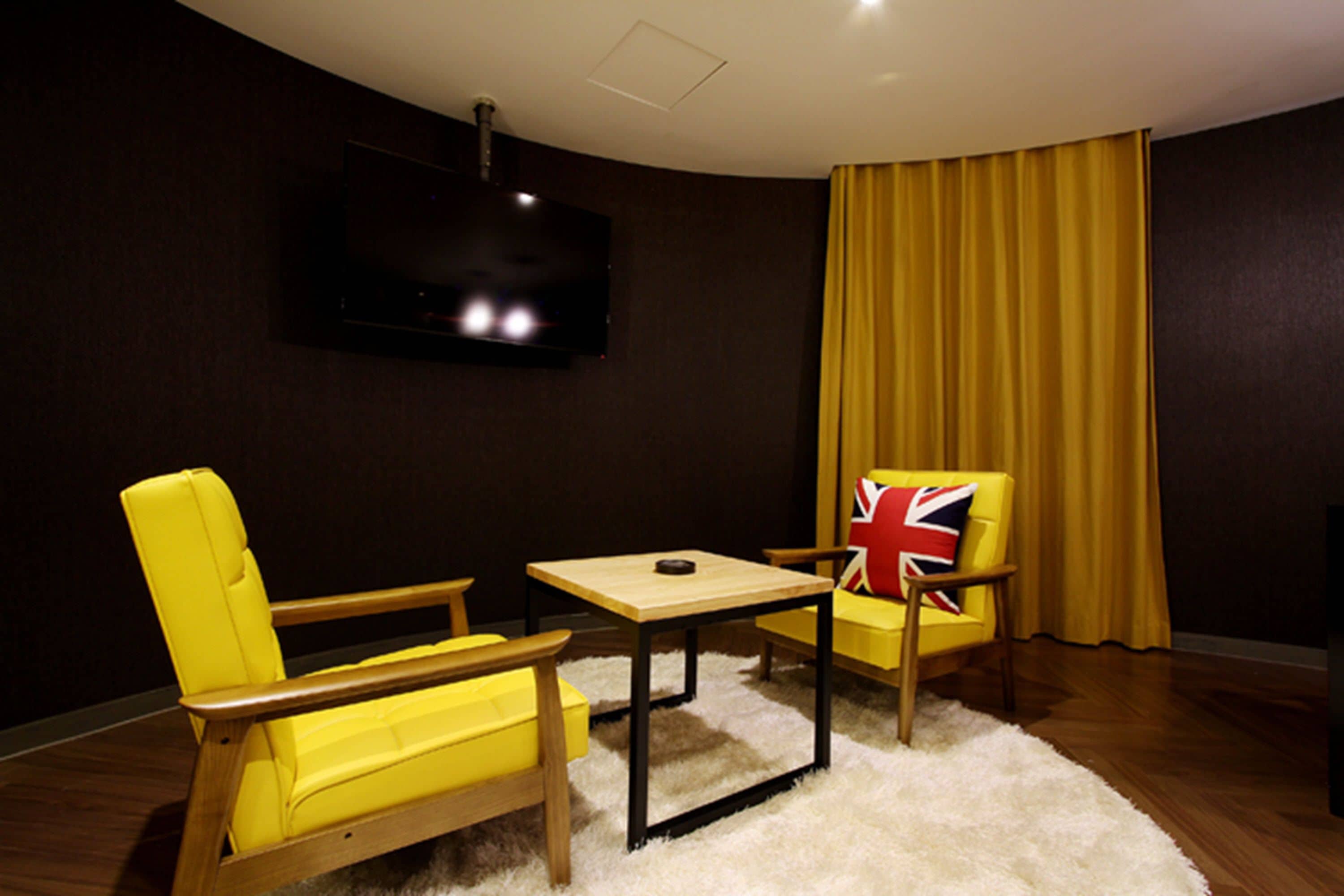 Een kamer met een wandgemonteerde flatscreen-tv, een gele fauteuil met een Union Jack kussen, een houten tafel en een geel gordijn.