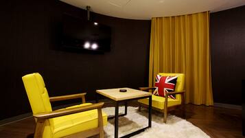 Een kamer met een wandgemonteerde flatscreen-tv, een gele fauteuil met een Union Jack kussen, een houten tafel en een geel gordijn.