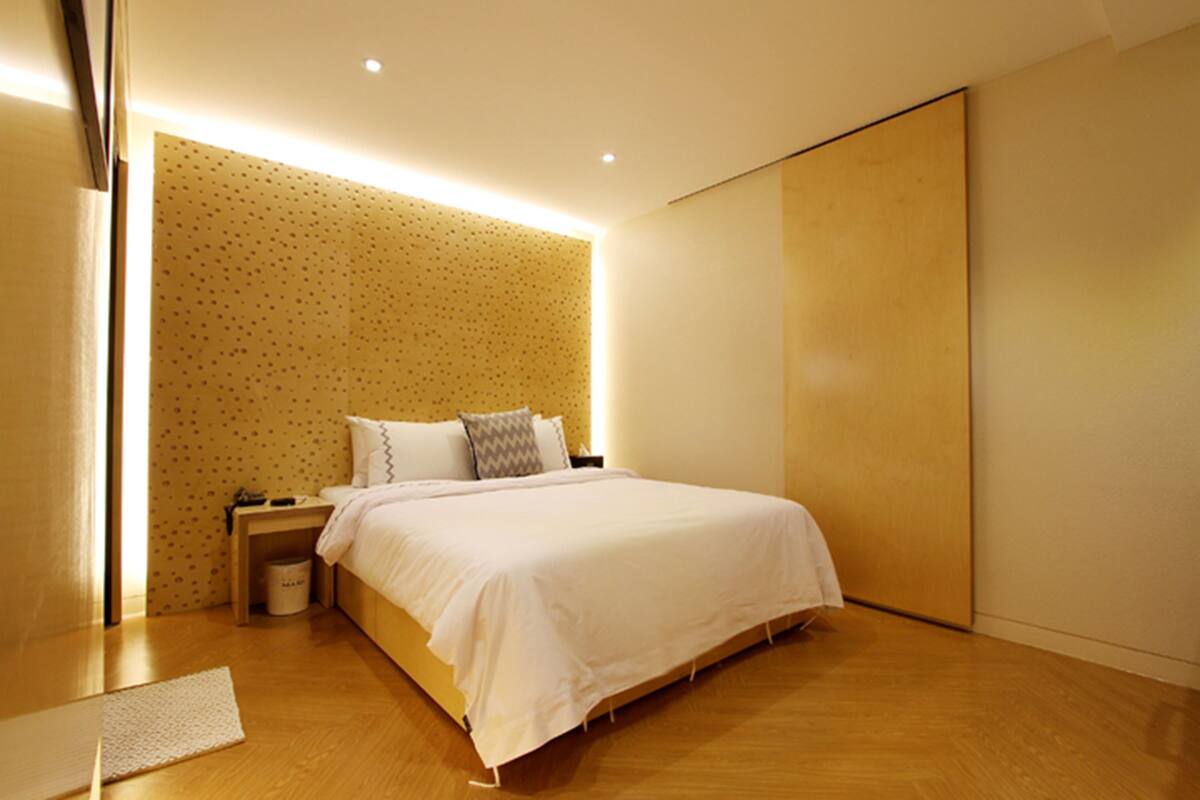 A modern bedroom with a large bed, a nightstand, a wall-mounted shelf, and a wooden floor.