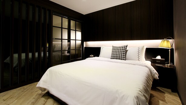 A modern bedroom with a large bed, dark wood paneling, a nightstand with a lamp, and a sliding glass door.
