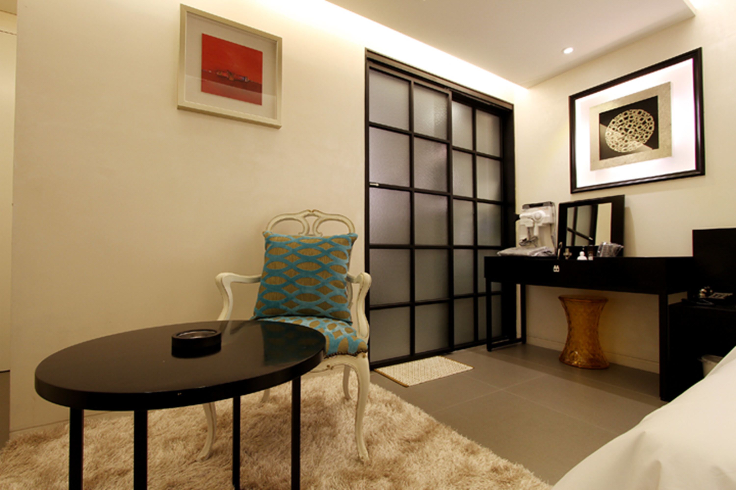 A hotel room with a round table, a chair with patterned cushions, a black desk with a mirror, and a framed artwork on the wall.
