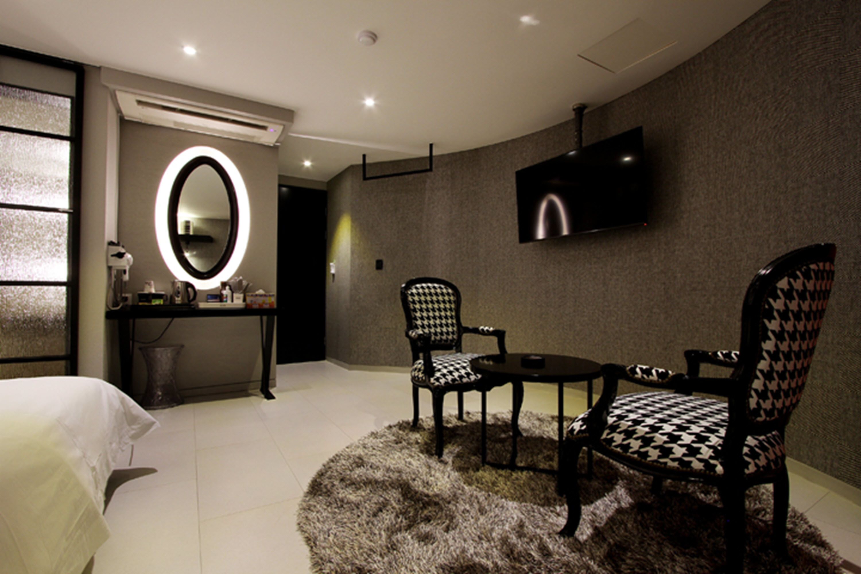 Quarto de hotel moderno com uma cama grande, uma mesa redonda com duas cadeiras, uma TV fixada na parede e um espelho com luz acima.
