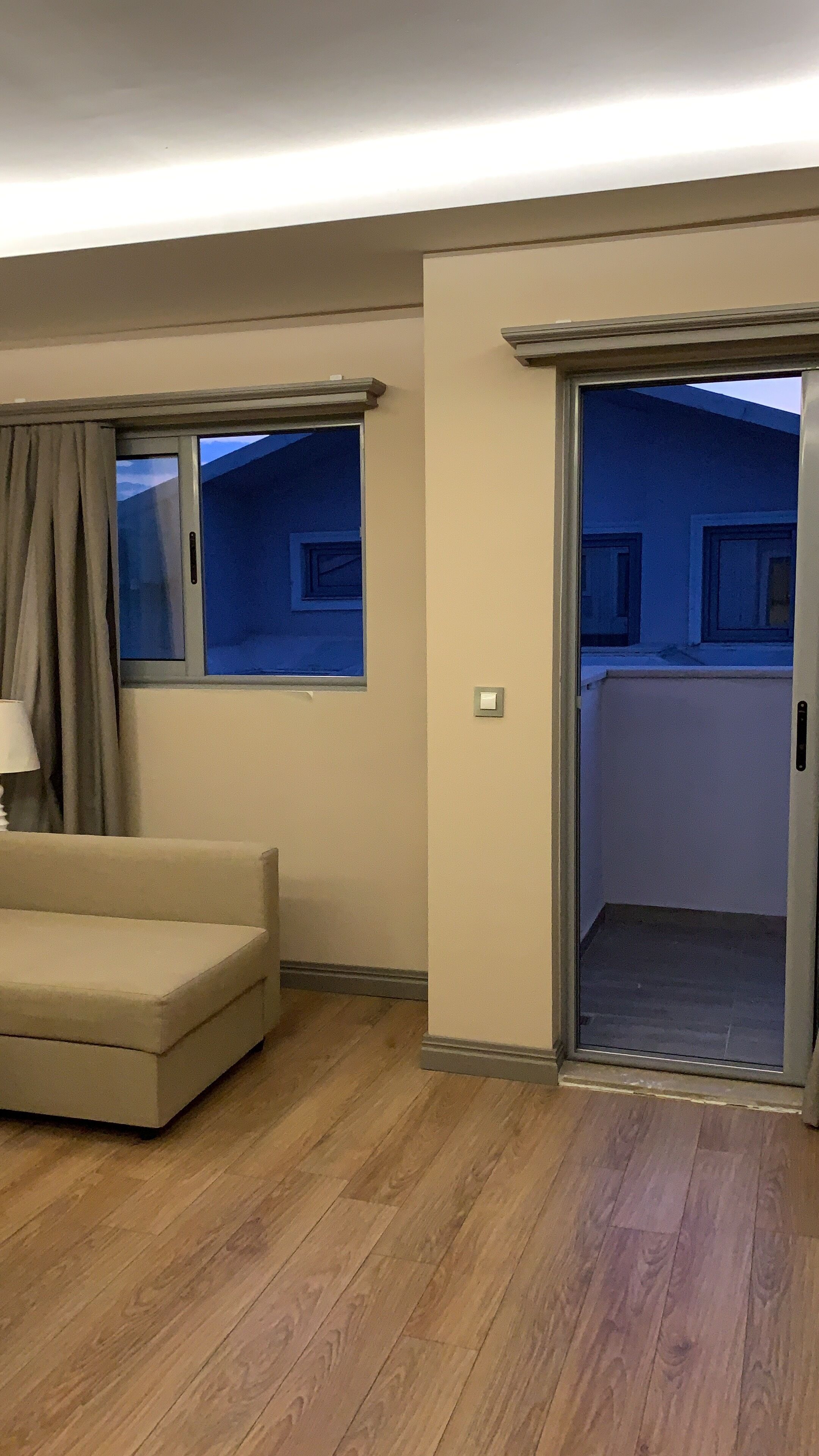 A room with a beige sofa, a window with curtains, and a door leading to another room.