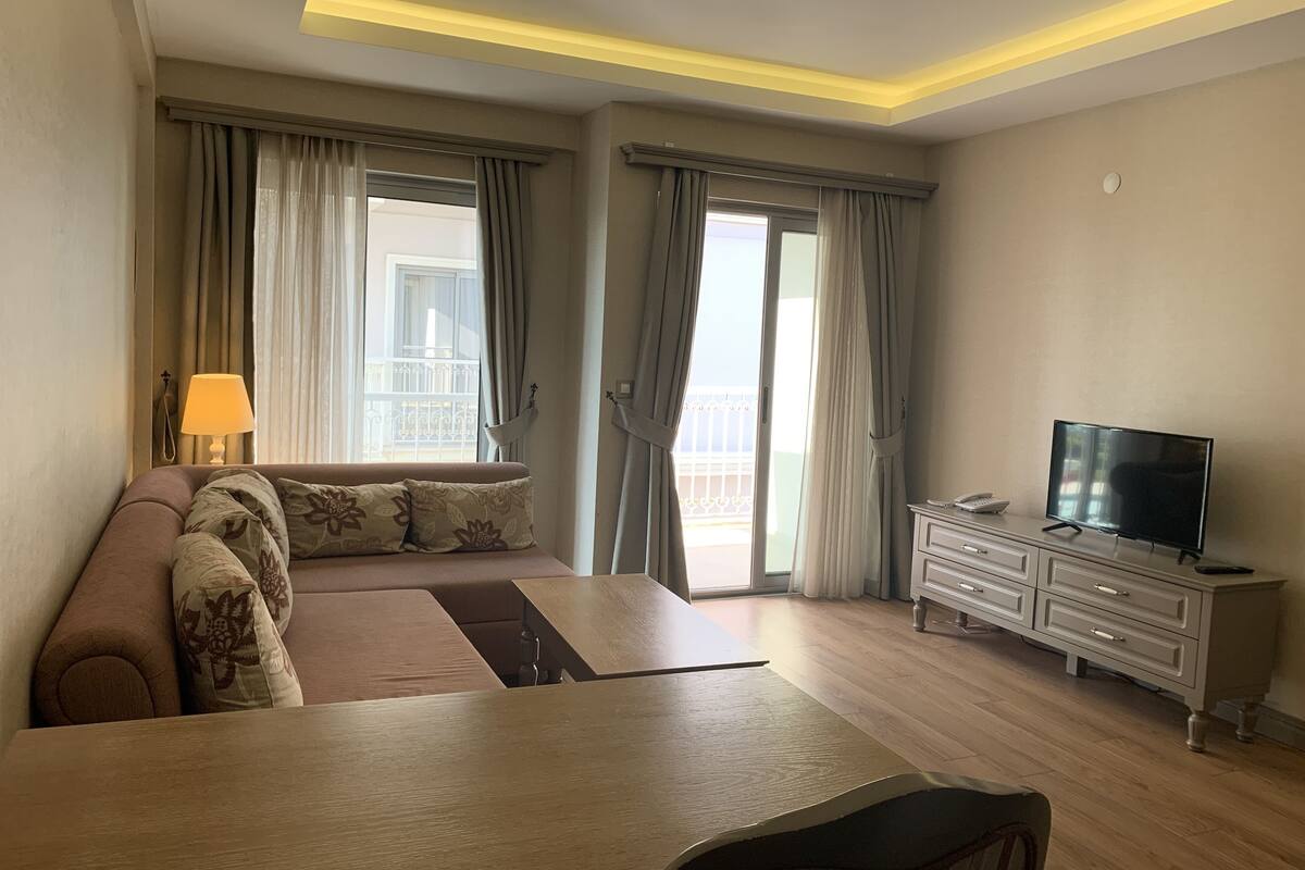 Une chambre d’hôtel avec un canapé, un téléviseur posé sur une commode, une table et un balcon avec des rideaux.