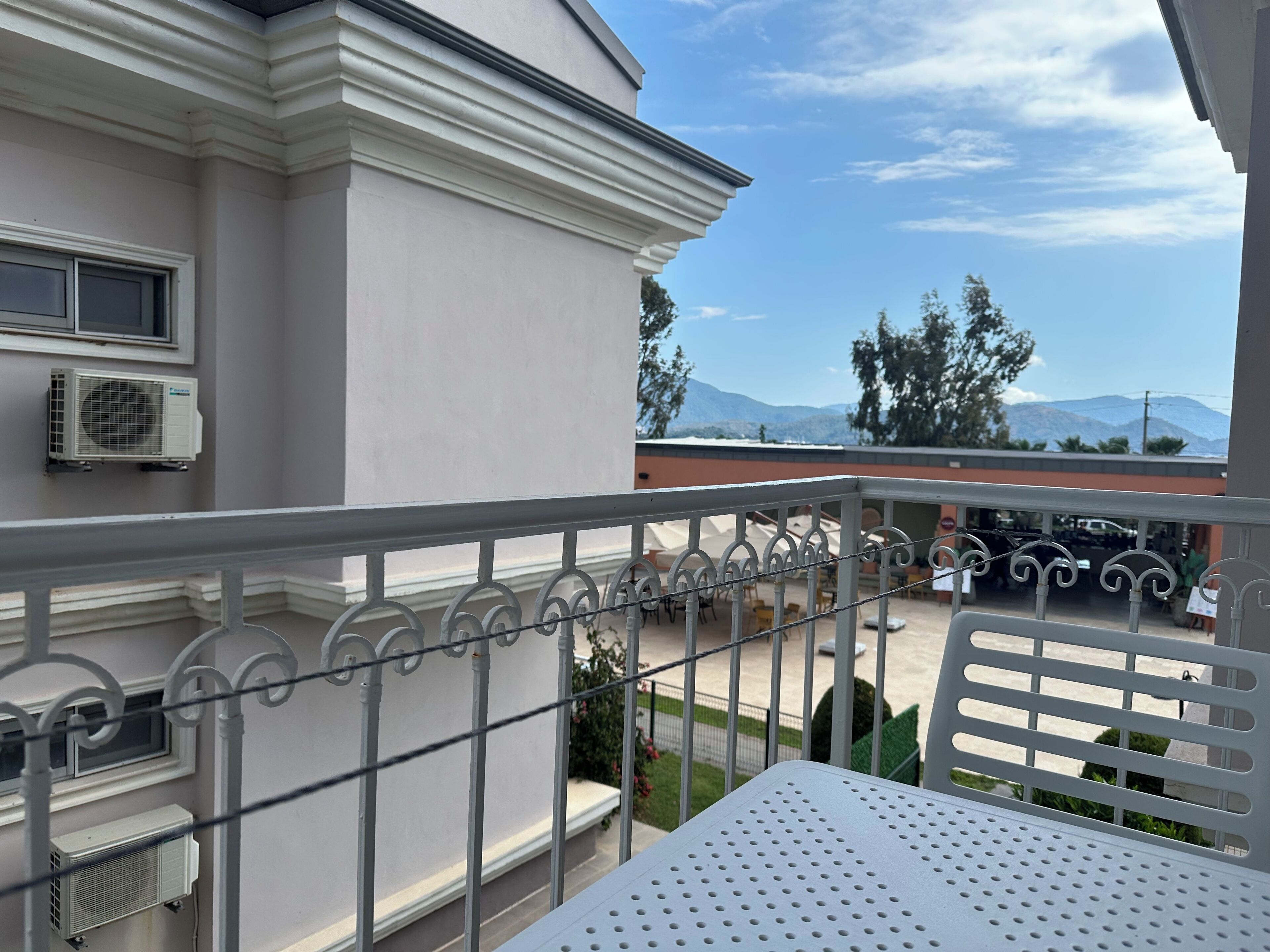 Ein Balkon mit Metallgeländer, eine Klimaanlage an der Wand und im Hintergrund ein Gebäude sowie Berge.