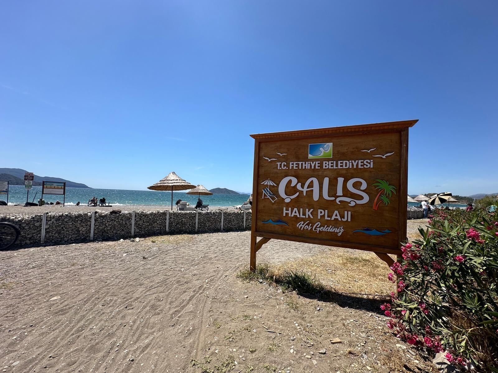 A beach sign with 'CALIS HALK PLAJI' and 'T.C. FETHIYE BELEDIYESI' text, surrounded by beach umbrellas and a stone wall.