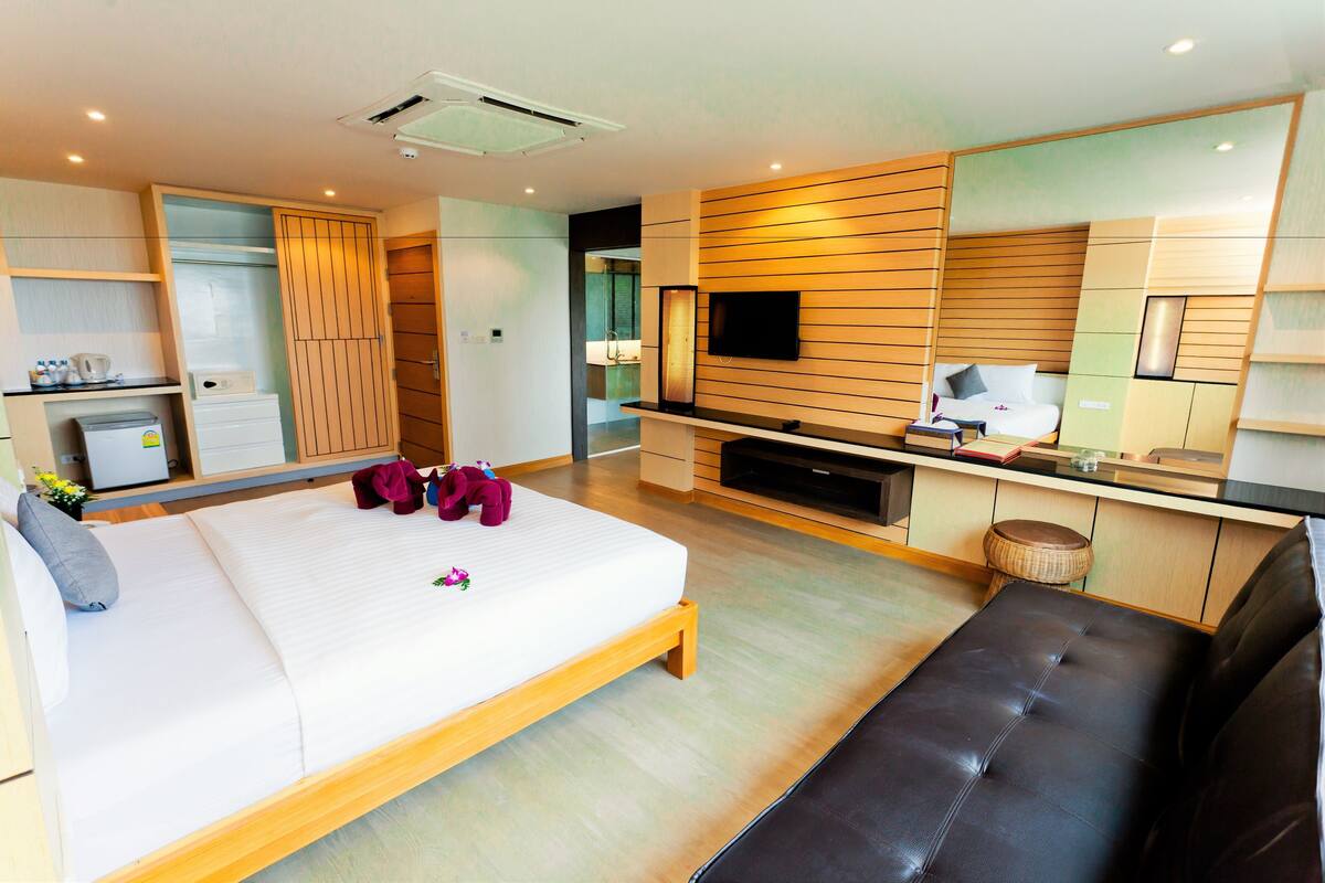 A modern hotel room with a large bed, a flat-screen TV, a wooden bench, and a kitchenette area.