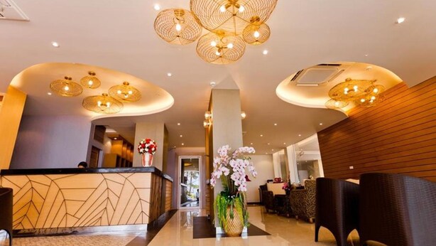 Um hall de hotel moderno com balcão de recepção de padrão geométrico, cadeiras de vime estilosas e um arranjo floral central.