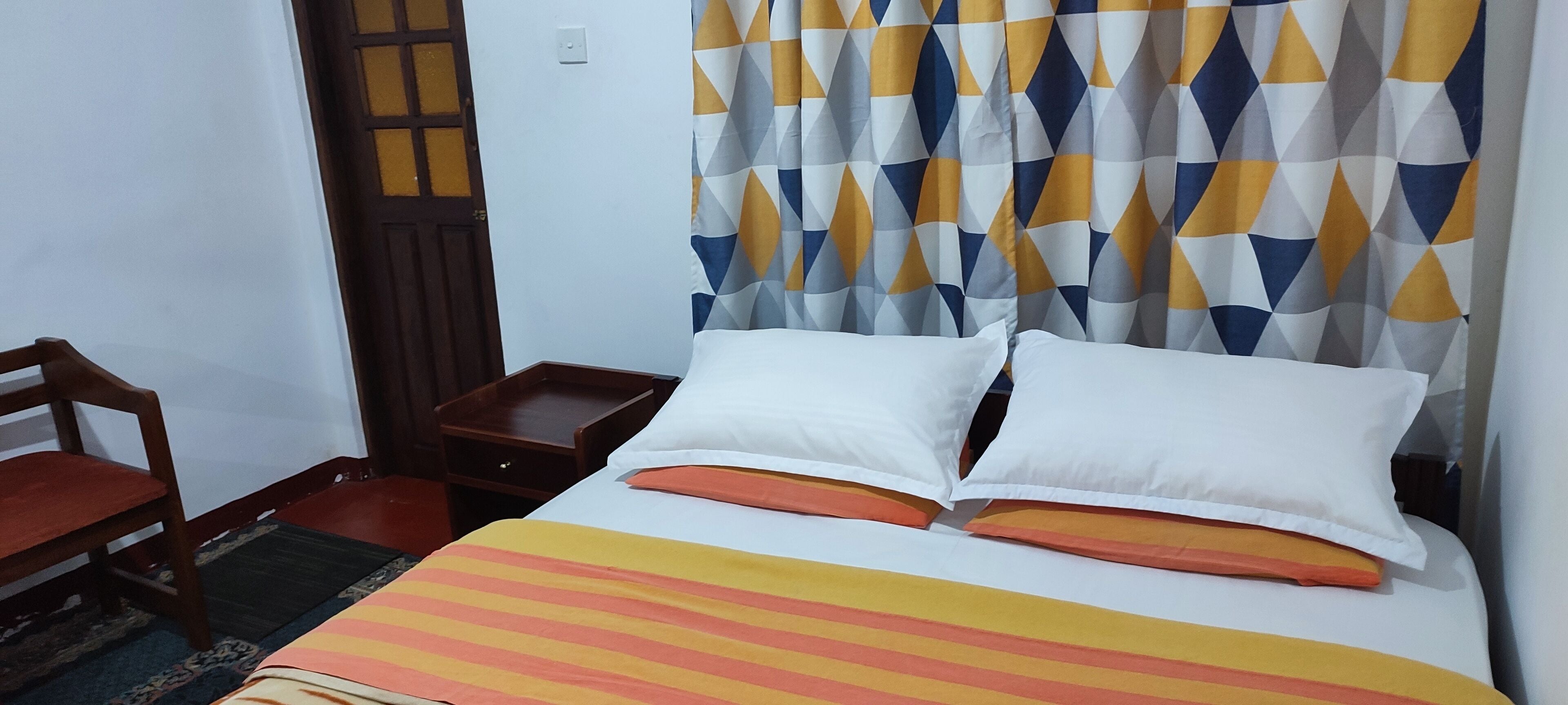 A hotel room with a bed, a wooden chair, a nightstand, and a patterned curtain.