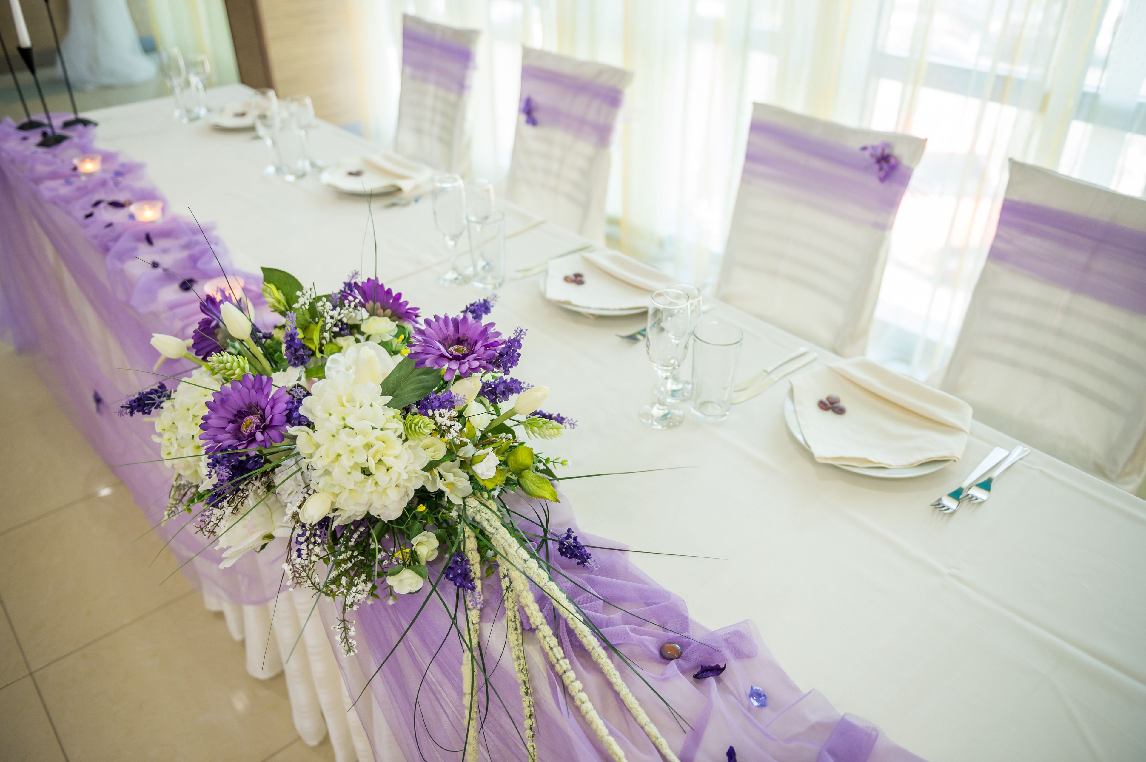 A long dining table with white tablecloths, purple floral centerpieces, and white napkins.