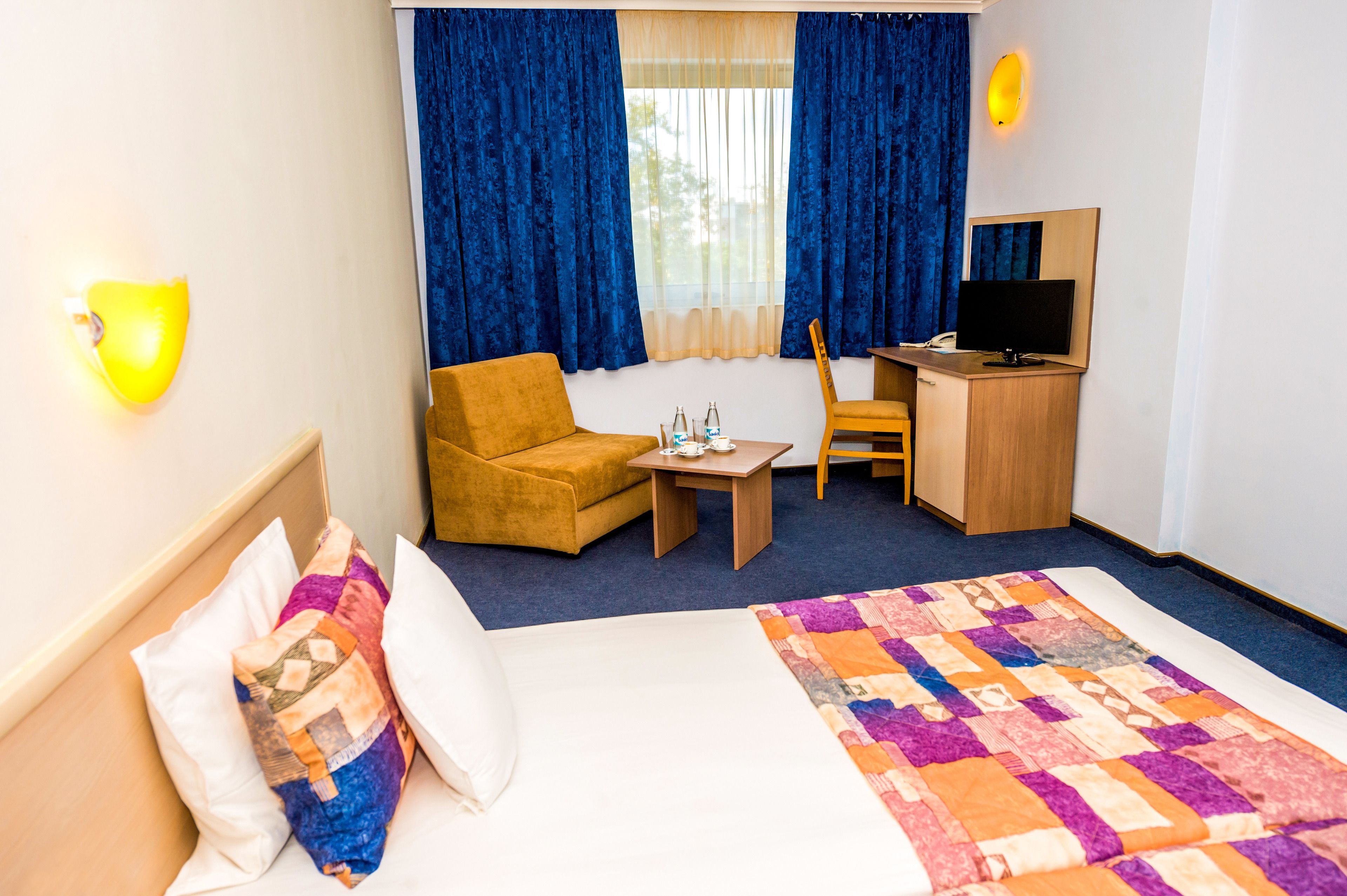 A hotel room with a bed, a sofa, a desk with a computer, a small table with bottles, and a window with curtains.
