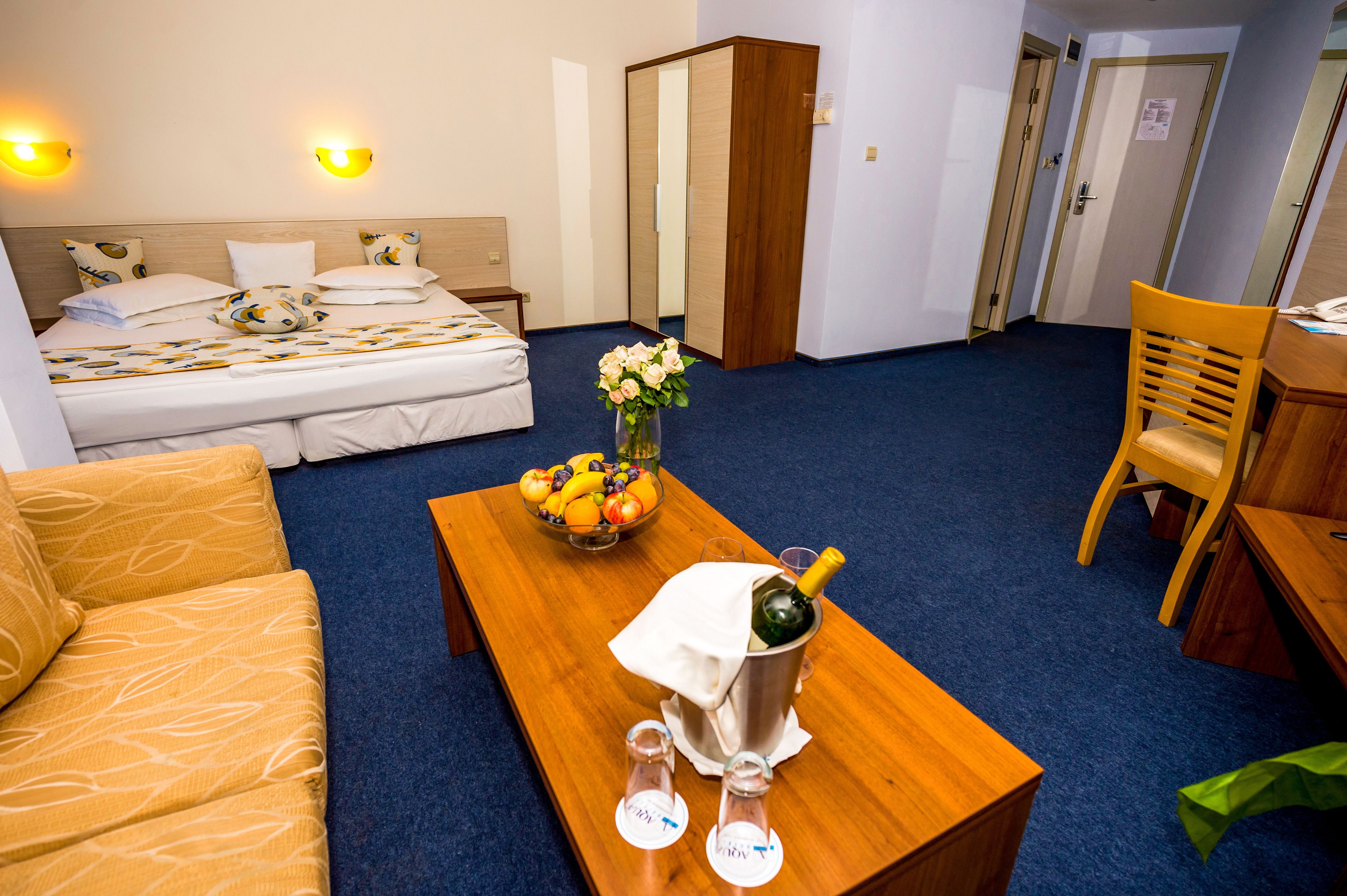 Ein Hotelzimmer mit einem Bett, einem Holztisch mit Früchten und einer Champagnerflasche, einem Stuhl und einem kleinen Tisch mit Gläsern.
