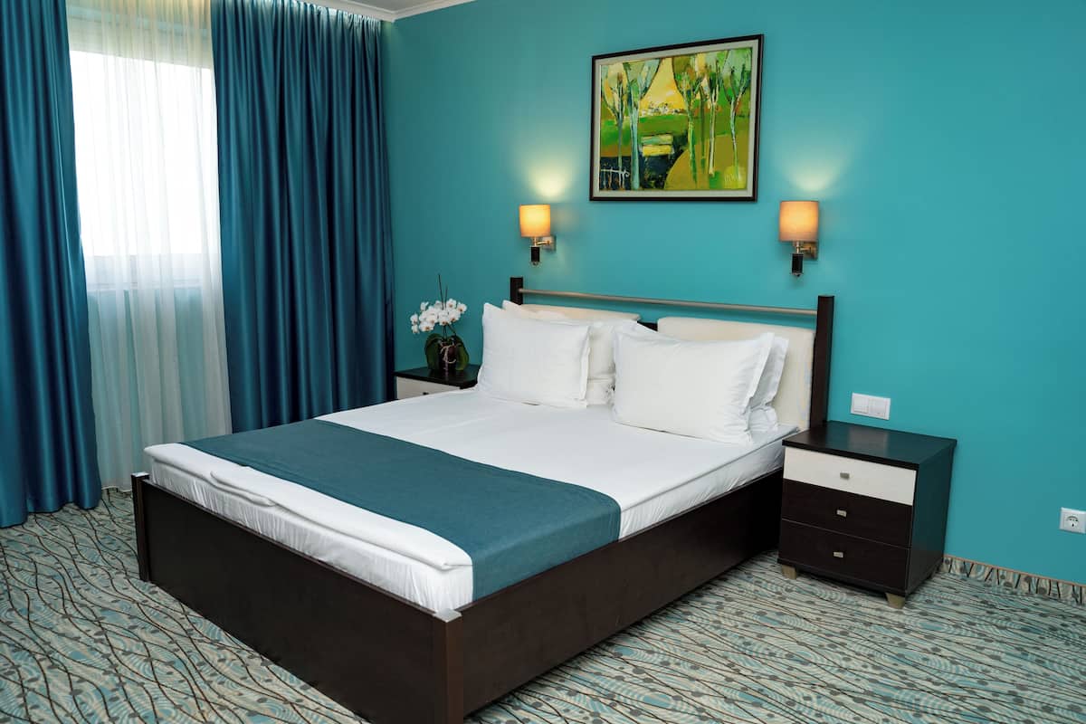 A hotel room with a large bed, two bedside tables, a nightstand, a painting, and a window with curtains.