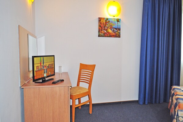 Habitación de hotel con escritorio, una silla, televisor, un cuadro en la pared y ventana con cortinas azules.