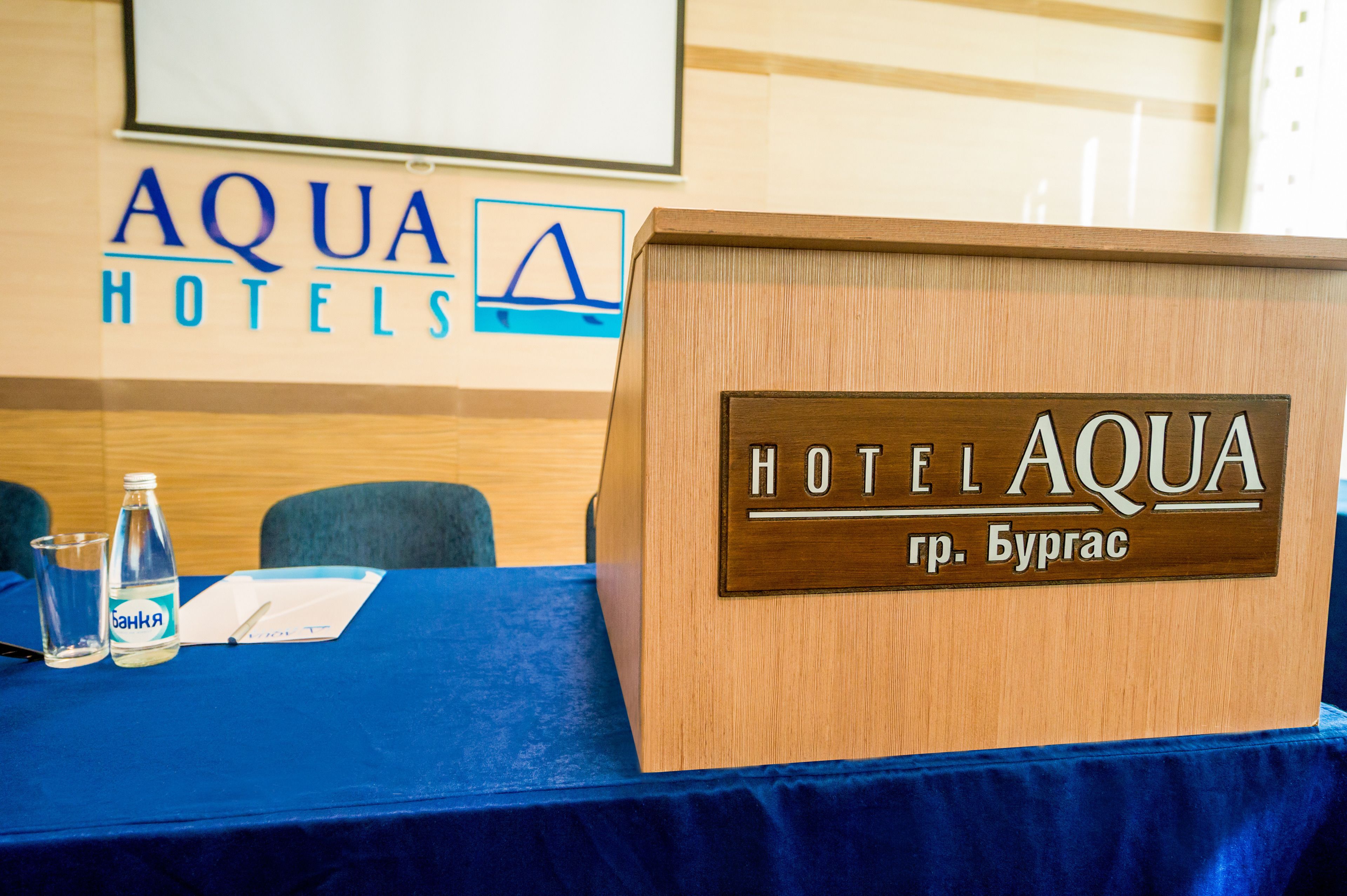 Un atril de madera con un cartel que dice "HOTEL AQUA" y "Hotel Aqua" en inglés, sobre una mesa azul con una botella de agua y vasos.