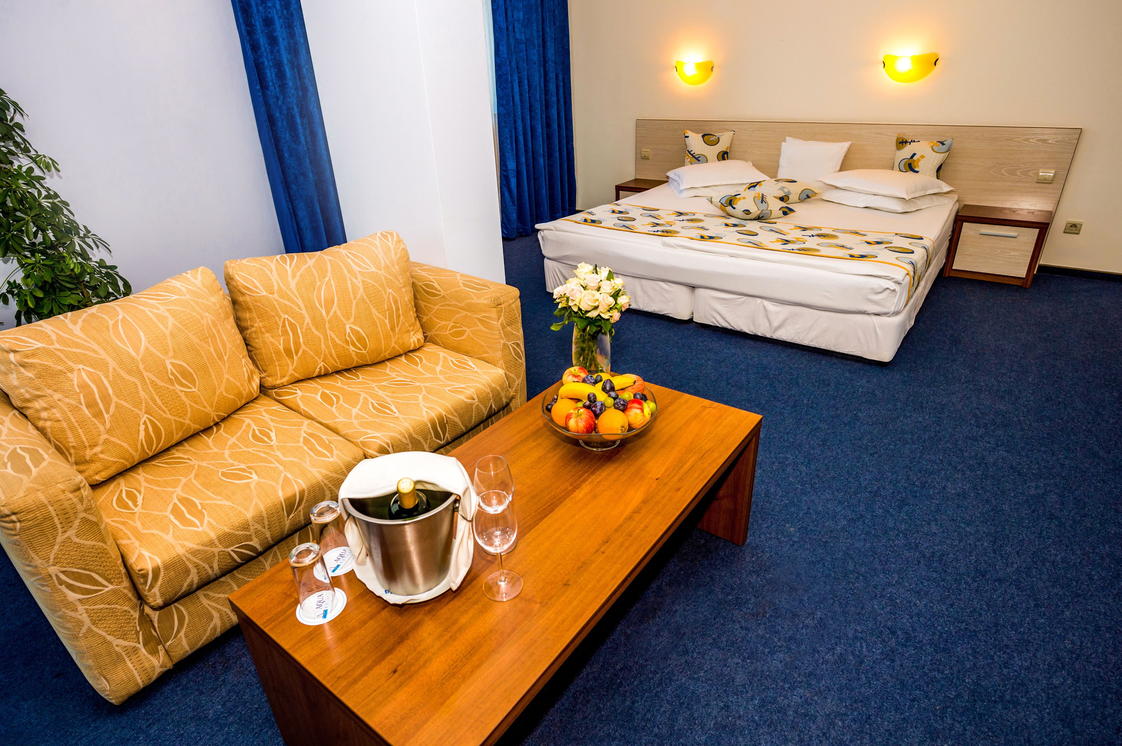 Quarto de hotel com sofá, cama com roupa de cama estampada, mesa de madeira com uma fruteira e champanhe, e duas arandelas.