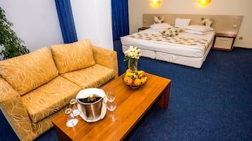 Quarto de hotel com sofá, cama com roupa de cama estampada, mesa de madeira com uma fruteira e champanhe, e duas arandelas.